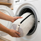 Twee handen schuiven de dichtgeritste crèmewitte waszak in de open trommel van een witte wasmachine