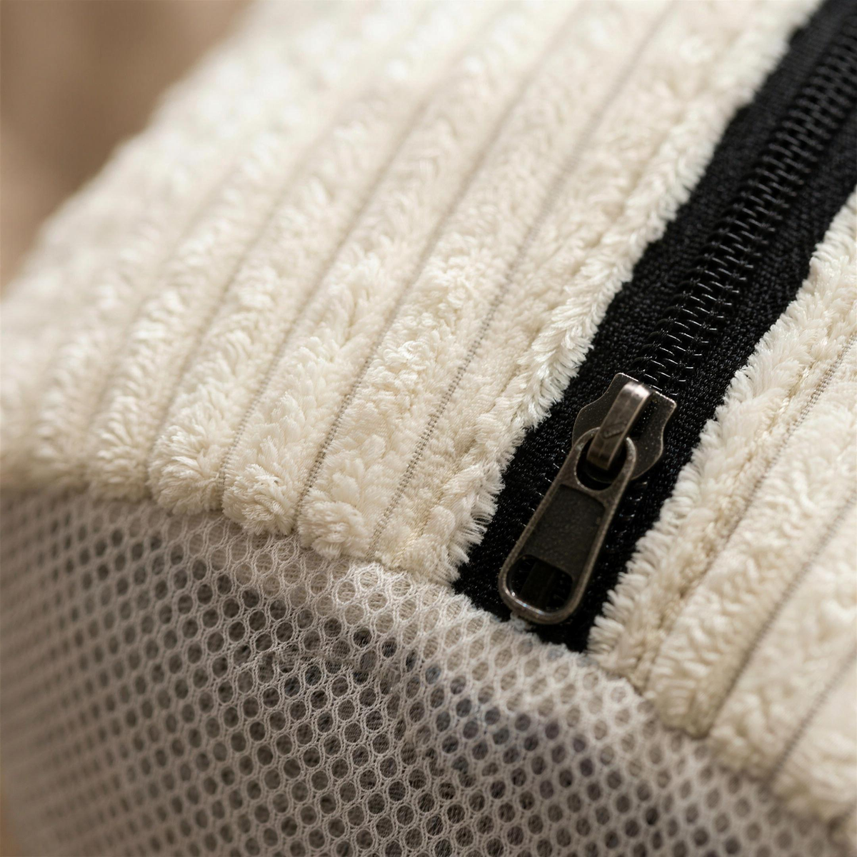 Macro-opname van de stof: dikke crèmewitte chenille ribben naast de zwarte rits met metalen schuiver en de rand waar het ribweefsel overgaat in het fijne grijze mesh