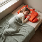 Slapend kind in een eenpersoons kinderbed met het hoofd op het oranje vossenkussen, saliegroene mousseline deken over het bed en daglicht uit het zijraam