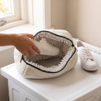 De waszak ligt half opengeritst op de wasmachine, een hand legt er een witte sneaker in; de binnenwanden zijn dicht bezet met witte chenille-slierten en onderin is het grijze meshpaneel zichtbaar, een tweede sneaker staat ernaast