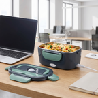 Lunchbox met warme pasta op een kantoorbureau naast een laptop, aangesloten op het stopcontact