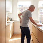 Man van rond de 60 staat in de keuken met zijn hand op zijn stijve onderrug
