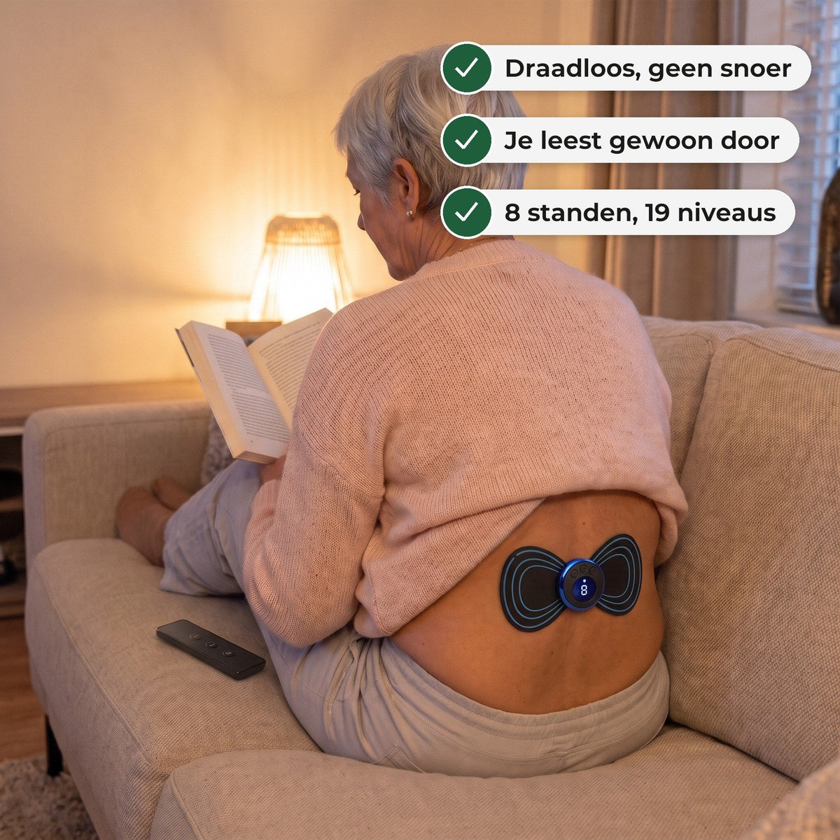 Vrouw van rond de 60 leest op de bank met de EMS-pad op haar onderrug, de afstandsbediening naast haar
