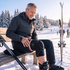 Man op een bankje aan de piste met de zwarte kniebrace om zijn knie, ski's naast hem in de sneeuw