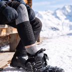 Korte zwarte scheenbeschermer met honingraatkussen over een grijze skisok, voet in een open skischoen op een terras in de sneeuw