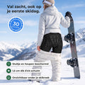 Snowboardster op de piste trekt haar witte skibroek iets omlaag en laat de zwarte valschort met stuitschild en heuppads zien, met drie uitleglabels en een zegel 30 dagen niet goed geld terug