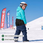 Lachend kind met skihelm en turquoise jas op een kinderpiste trekt een blauwe skibroek op over de zwarte valschort in kindermaat