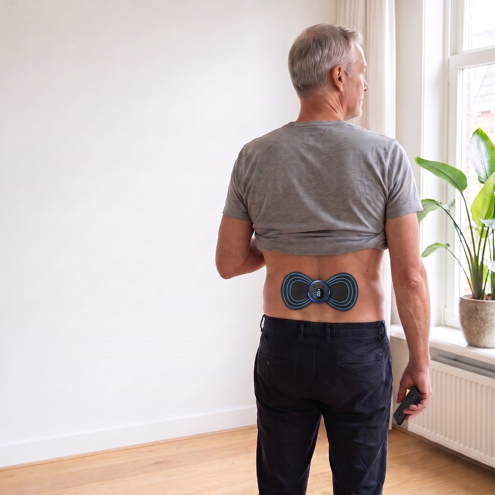 Man van rond de 60 staat in de woonkamer met de EMS-pad op zijn onderrug en de afstandsbediening in zijn hand